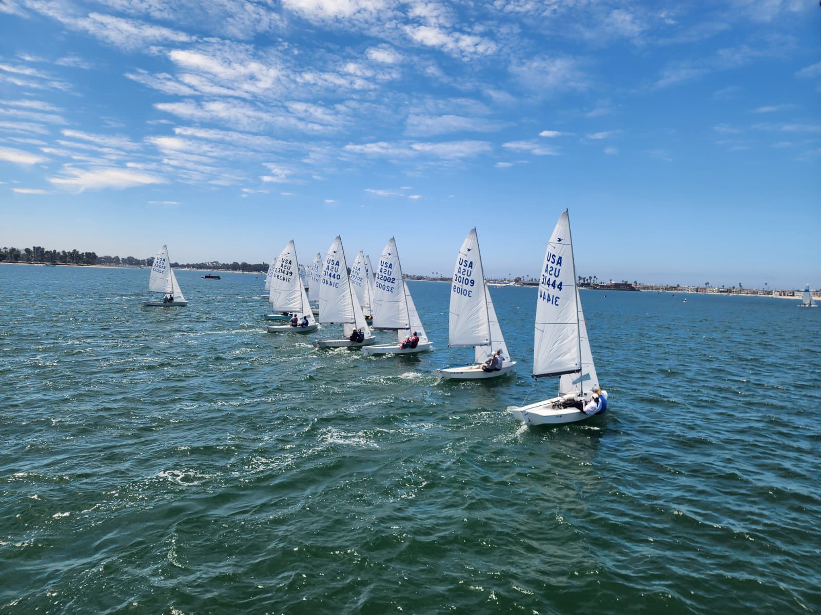 Seven small sailboats with white sails race across blue water under a partly cloudy sky. The boats are spaced in a line, sailing toward the right. The shoreline and some trees are visible in the background.