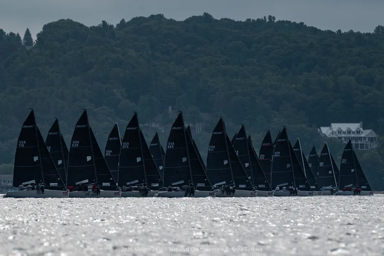 A line of black sailboats with numbers on their sails race on a lake, with forested hills and white houses in the background under a cloudy sky.