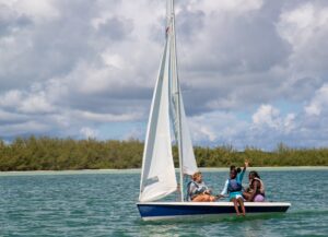 Three people sit on a small sailboat in calm blue water near a green shoreline under a partly cloudy sky; one person waves while others look forward.