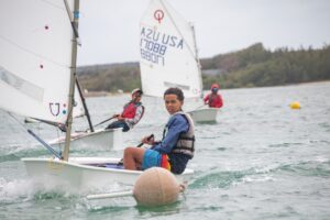 Three young sailors in small sailboats race on a lake, wearing life jackets. The closest sailor looks at the camera, with trees and a cloudy sky in the background. An orange buoy floats in the foreground.