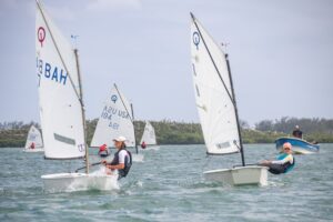 Four people are sailing small sailboats on open water, with two motorboats and land visible in the background. The sailors appear focused, and the weather is clear with light clouds.