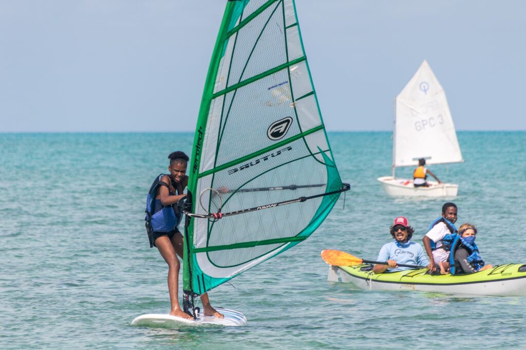 A person windsurfing near two people in a kayak and a small sailboat on a calm blue sea under a clear sky.