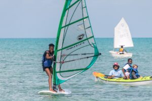 A person windsurfing near the shore with a green sail, while three people in a yellow kayak and another person sailing a small boat are visible in the background. The sea is calm and the sky is clear.