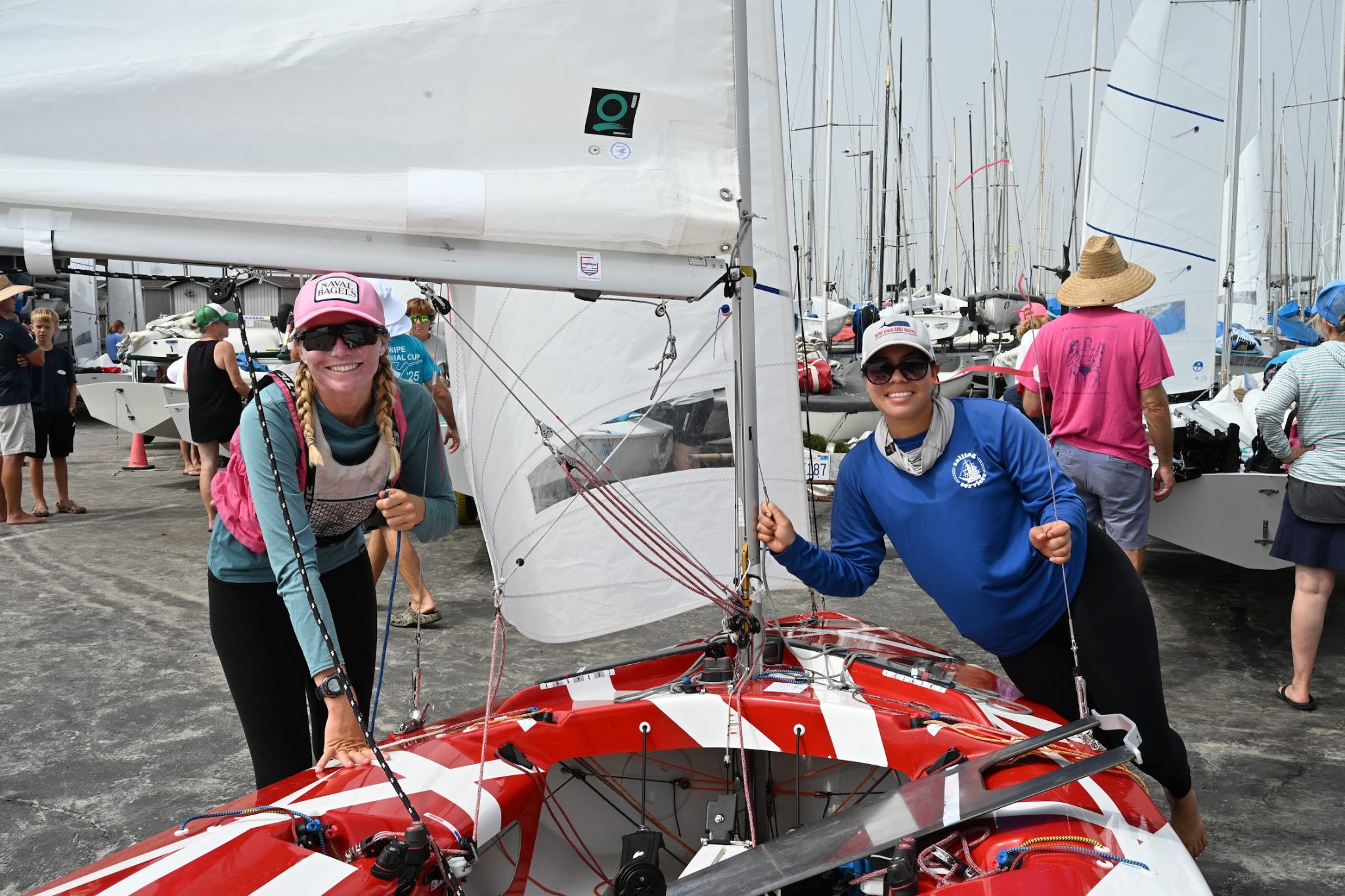 Two smiling women stand beside a red-and-white sailboat, holding its rigging. Sailboats and people are in the background at a marina. Both women wear hats and sunglasses.