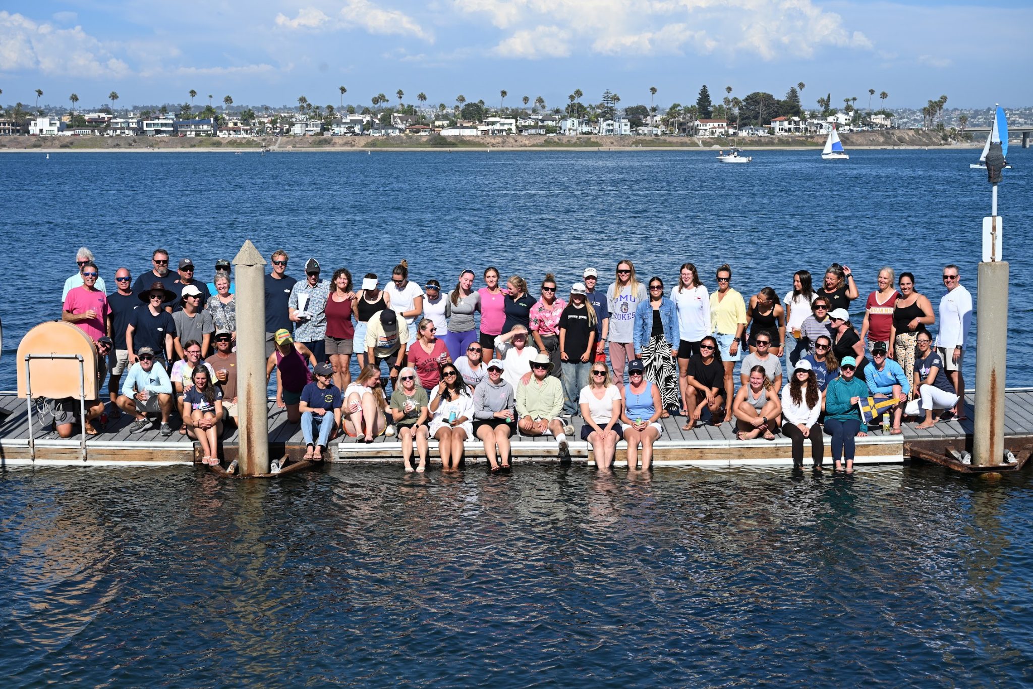 A large group of people poses together on a dock over calm water, with a coastal town, palm trees, and a sailboat visible in the background under a partly cloudy sky.