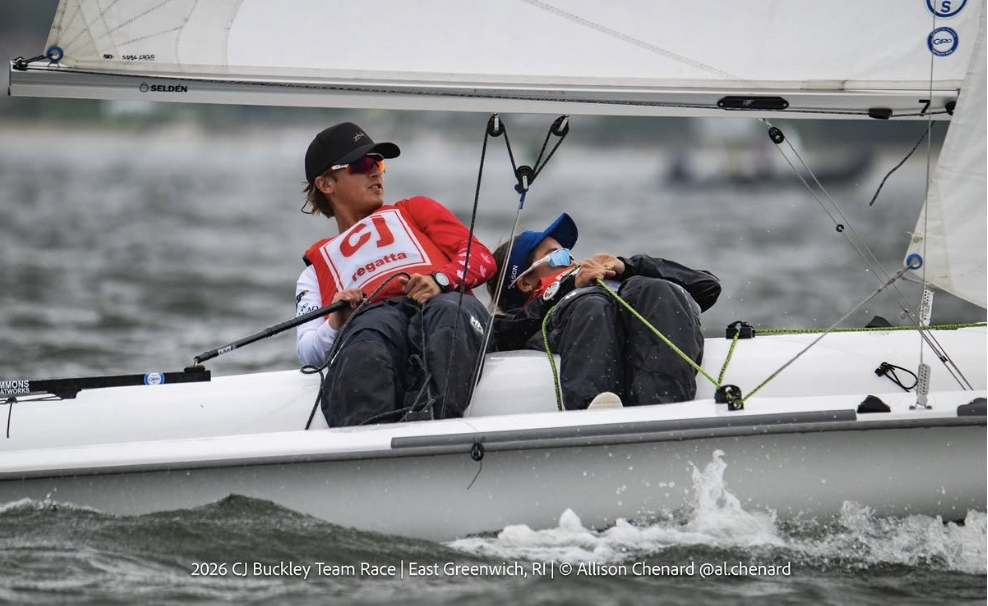 Two sailors wearing life jackets and caps lean out of a small sailboat during a race on choppy water. The boat’s sail is angled and both are focused on maneuvering in windy conditions. Text overlay indicates event details.