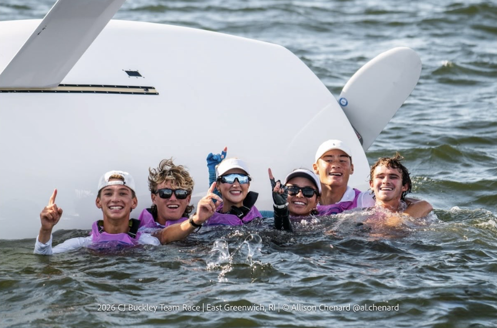 Six young people in life jackets smile and hold up one finger while standing in the water next to a capsized sailboat, celebrating together under sunny skies.