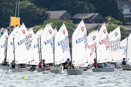 A group of young sailors competes in a race with small sailboats, each displaying a white sail marked “USA” and numbers. Houses and trees are visible in the background.