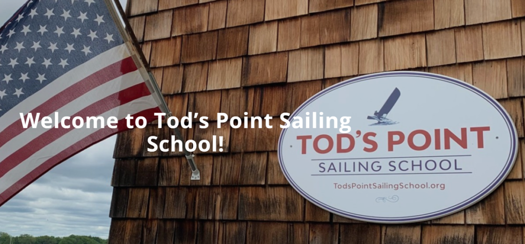 An American flag and a sign reading “TOD’S POINT SAILING SCHOOL” are mounted on a shingled wall. White text says, “Welcome to Tod’s Point Sailing School!”.