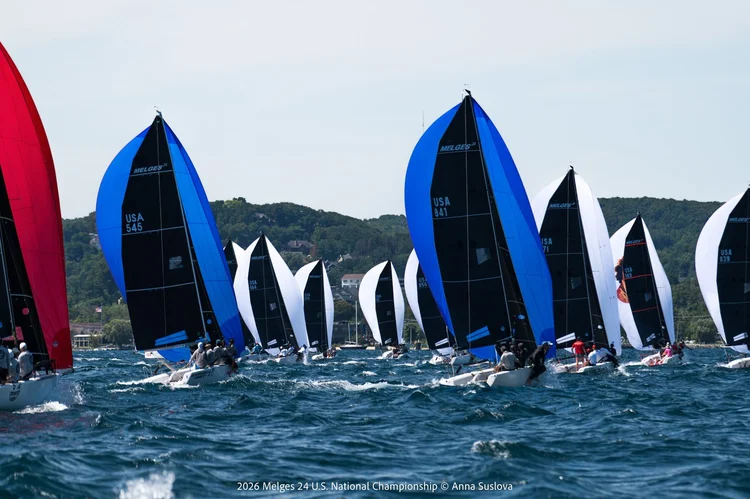 Sailboats with colorful spinnakers race on choppy blue water, with green hills and buildings in the background under a partly cloudy sky.