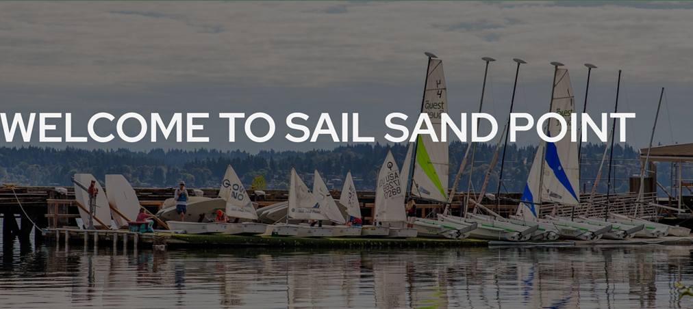Sailboats are docked by a wooden pier on calm water, with “WELCOME TO SAIL SAND POINT” in large white text across the image. Trees and clouds are visible in the background.