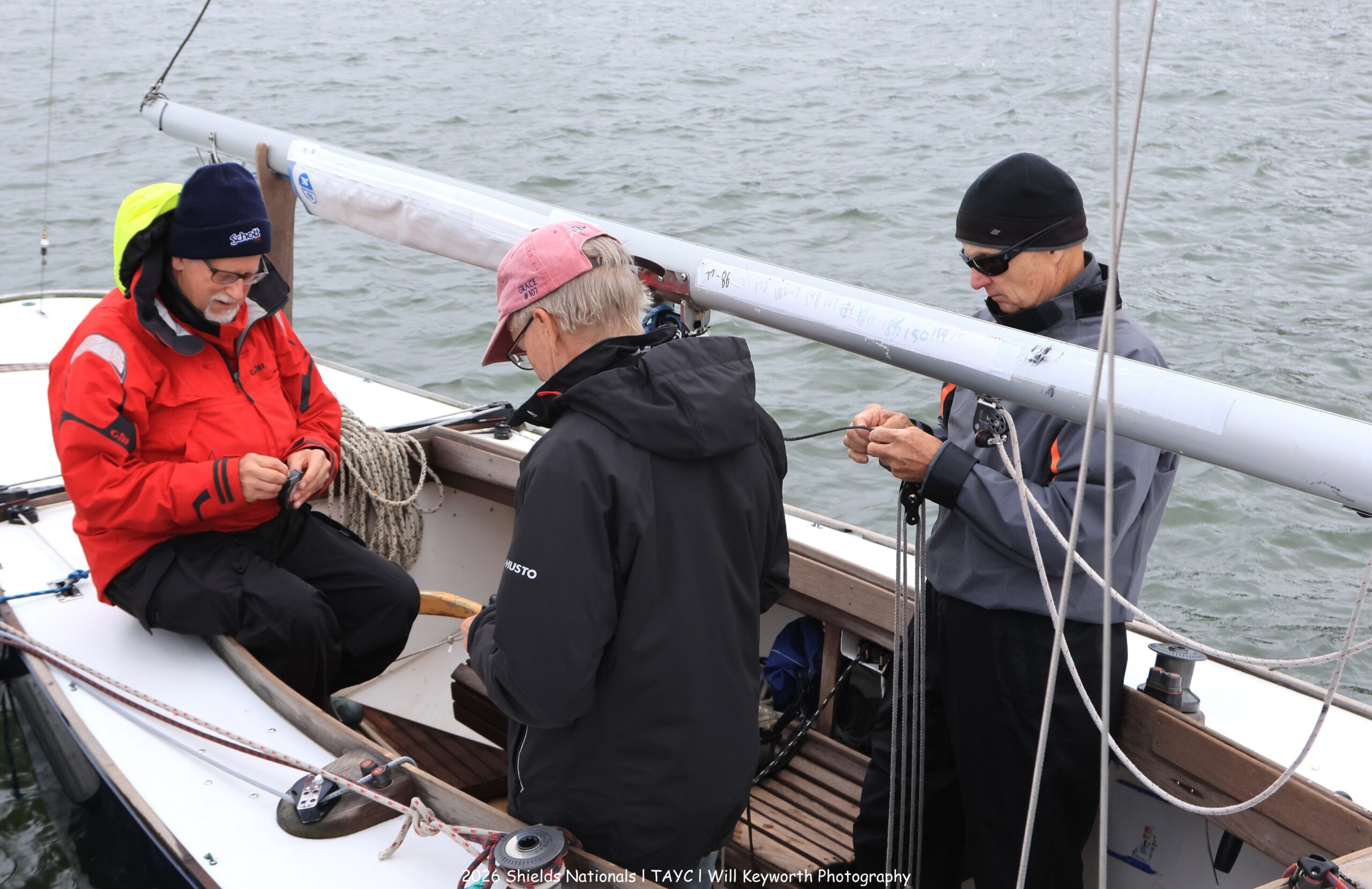 Three people in jackets prepare equipment on a small sailboat docked on the water, focusing on ropes and gear. The weather appears cool and overcast.