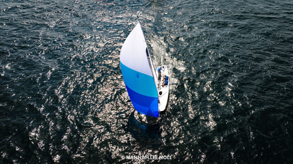 A small sailboat with a blue and white striped sail glides across dark, rippling water, sunlight reflecting off the waves.
