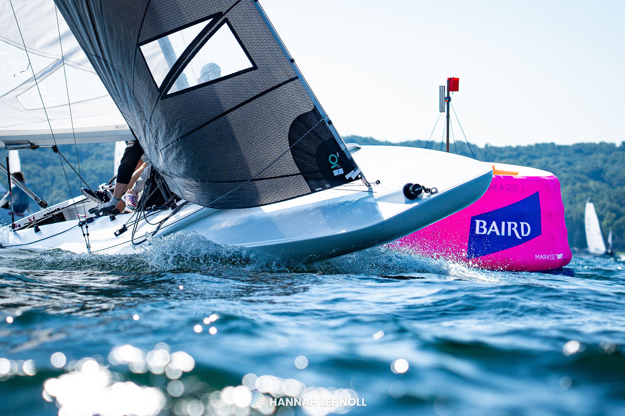 A sailboat races around a bright pink buoy on a sunny day, with water splashing near the boat and trees visible in the background.