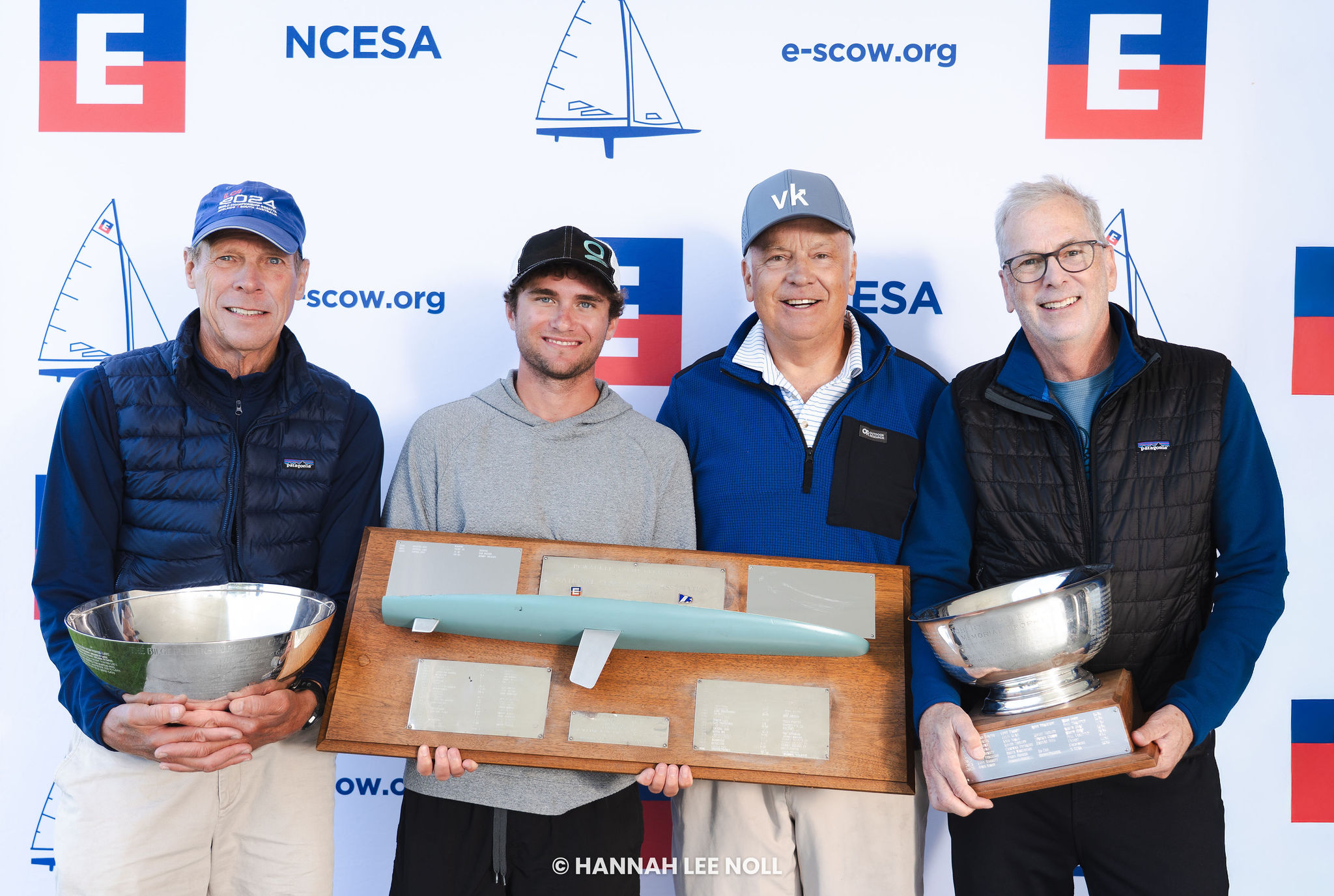 Four men stand in front of a sailing-themed backdrop, smiling and holding trophies and a model sailboat on a plaque, suggesting a sailing competition award ceremony.