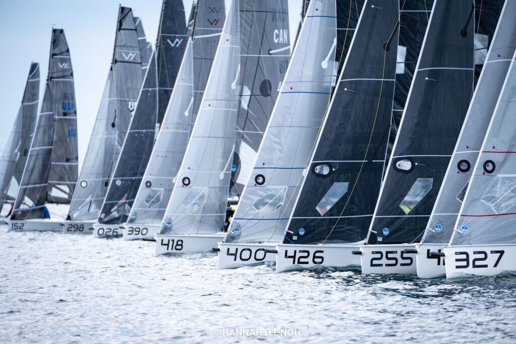 A row of numbered sailboats lined up on calm water, their sails unfurled and ready for a race under a partly cloudy sky.