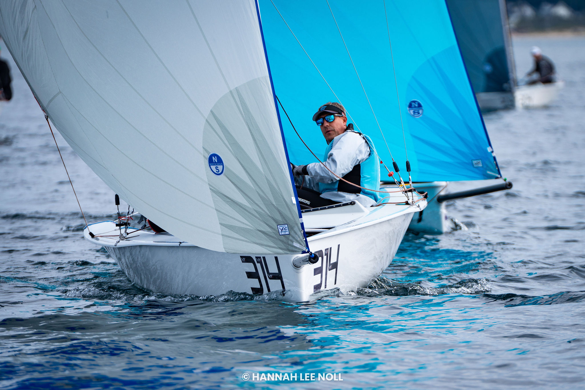 A sailor in sunglasses steers a white sailboat with the number 34, blue sails filled with wind, gliding over calm water during a sailing race. Other boats are visible in the background.