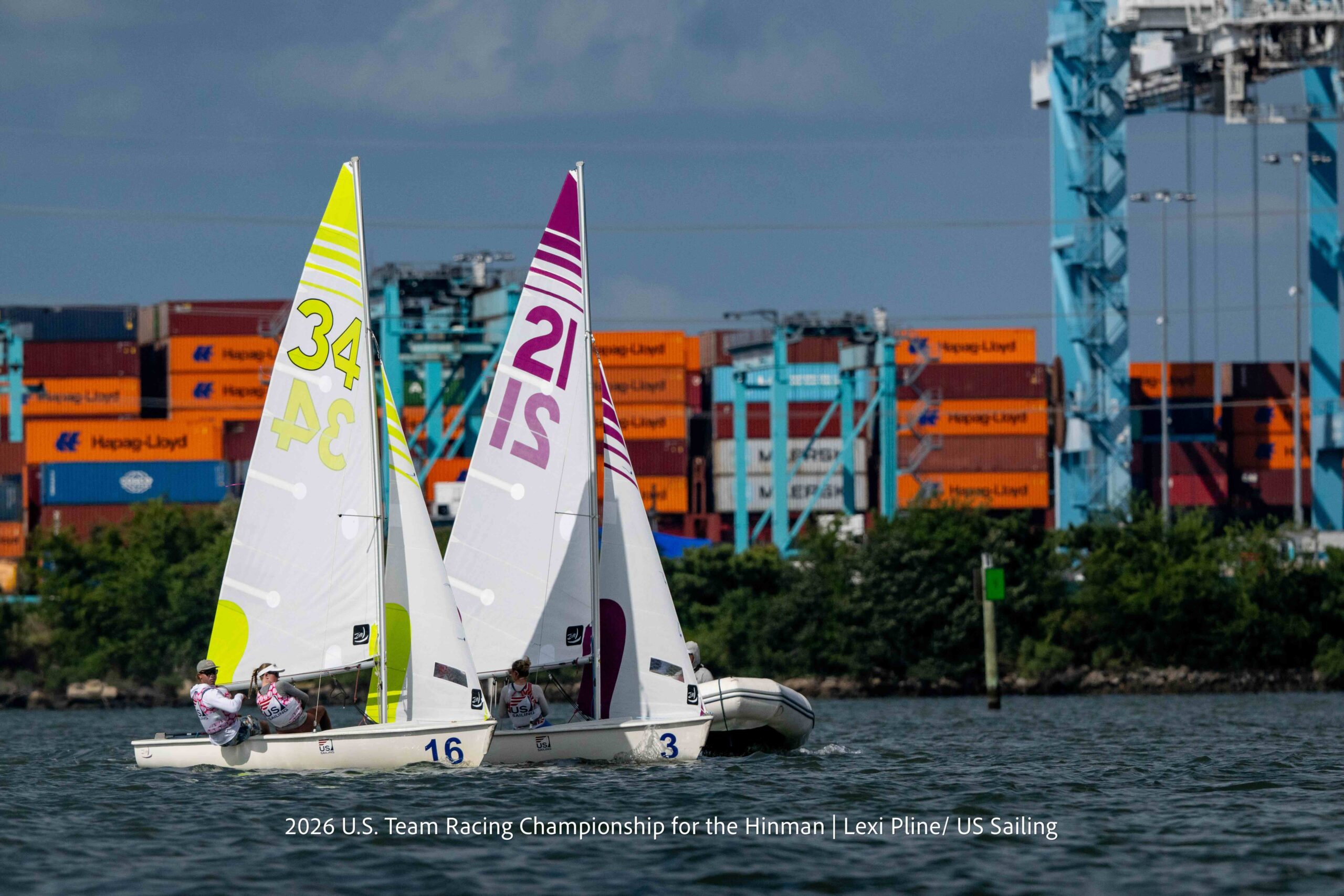 Two small sailboats with numbers 34 and 21 race side by side on water, with people onboard. Shipping containers and cranes are visible in the background under a cloudy sky.