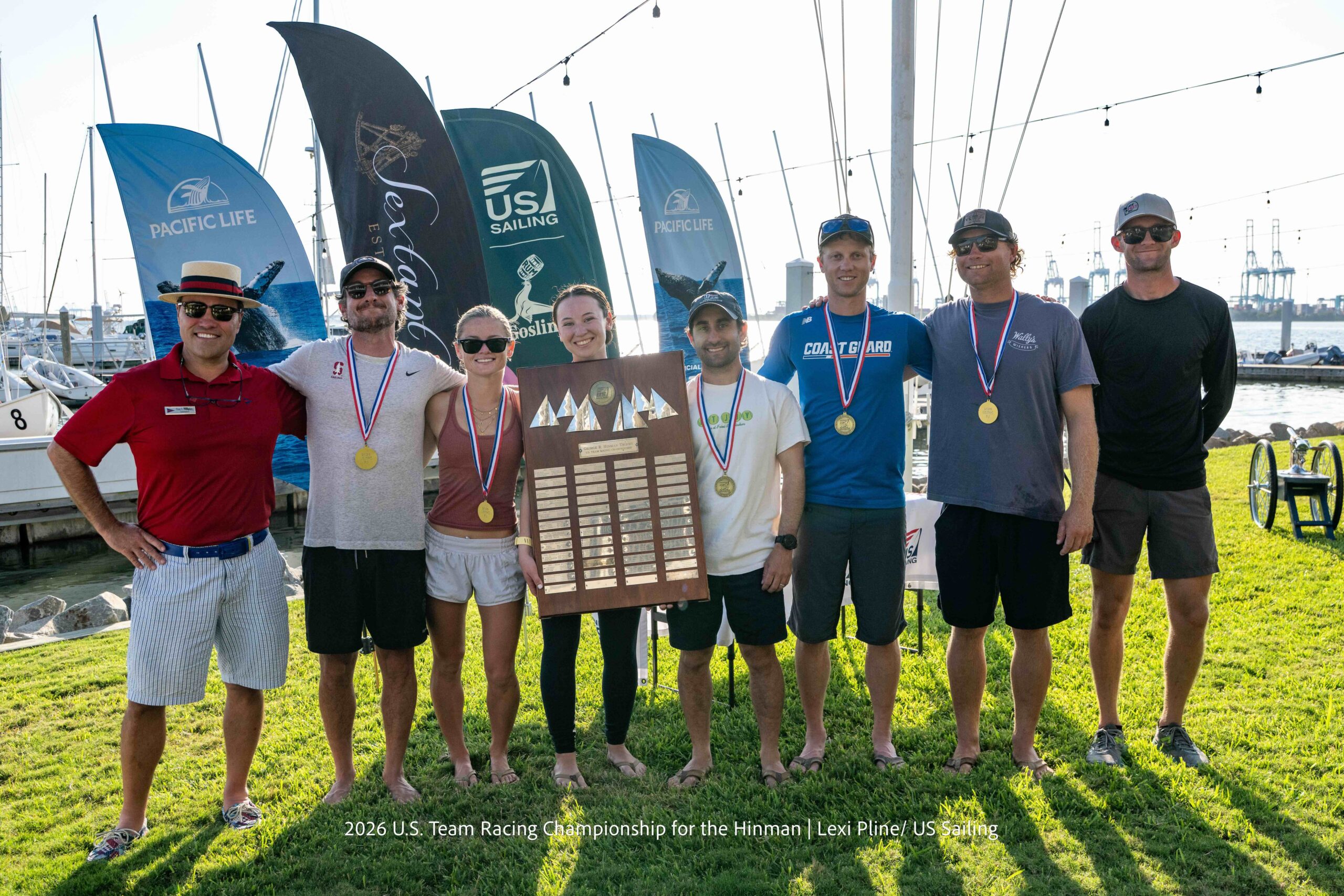 Eight people stand on grass by sailboats, smiling and wearing medals; four people in the center hold a large wooden trophy. Banners and masts are visible in the background on a sunny day.