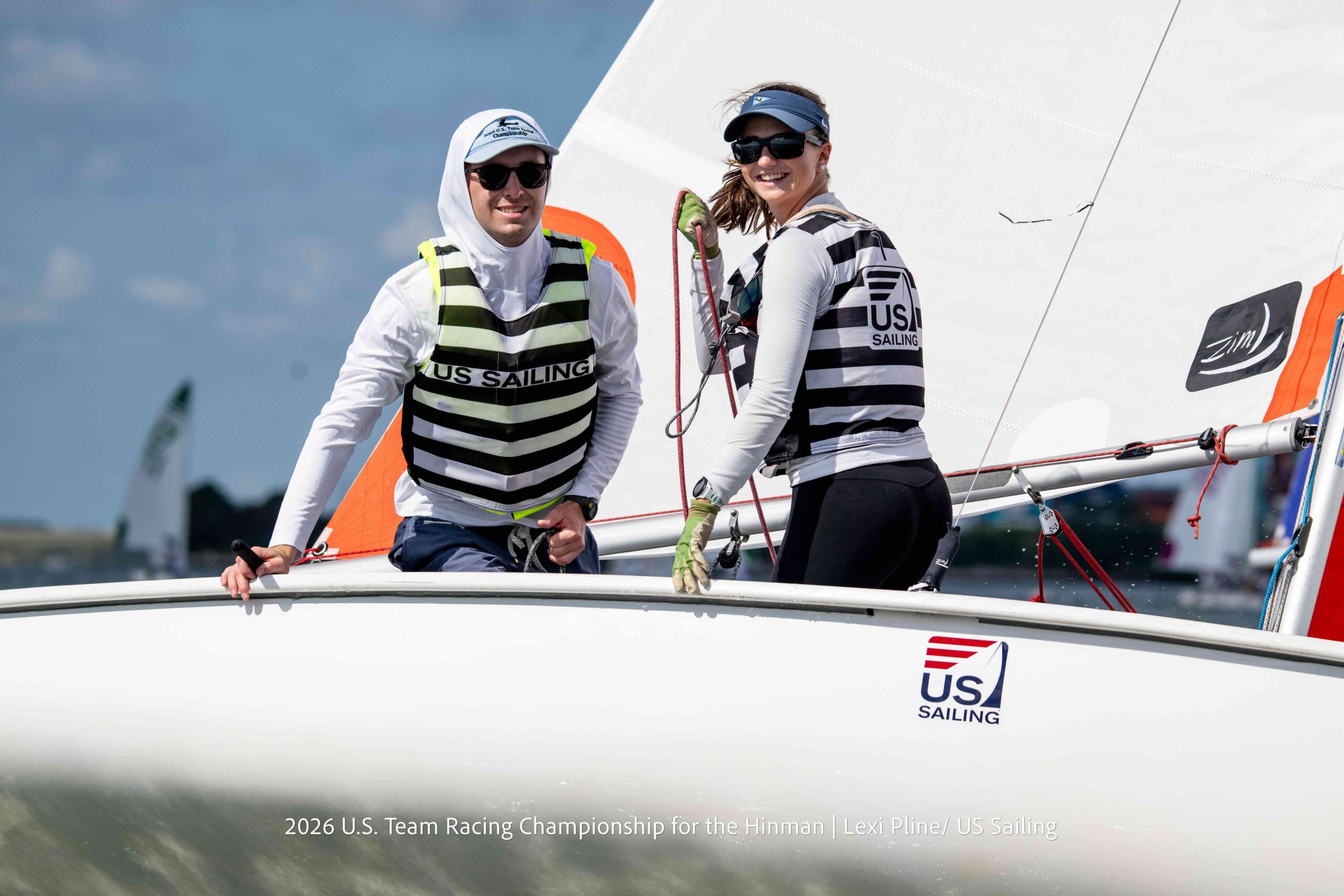 Two sailors in life vests and sunglasses smile on a sailboat with “US Sailing” logos, holding ropes and steering during a race.