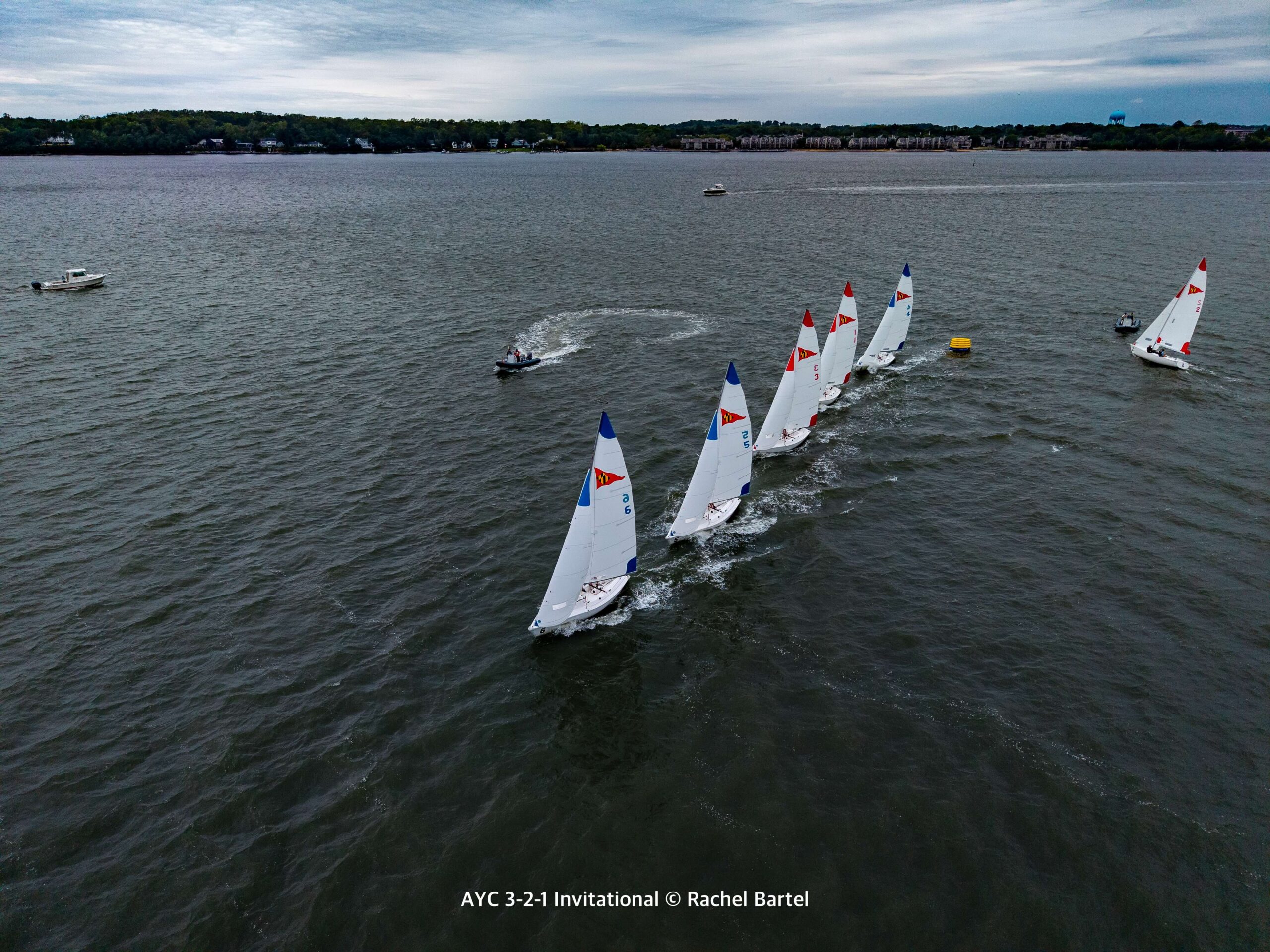 Aerial view of seven small sailboats racing on a lake, heading toward a yellow buoy, with escort boats nearby and a tree-lined shore in the background.