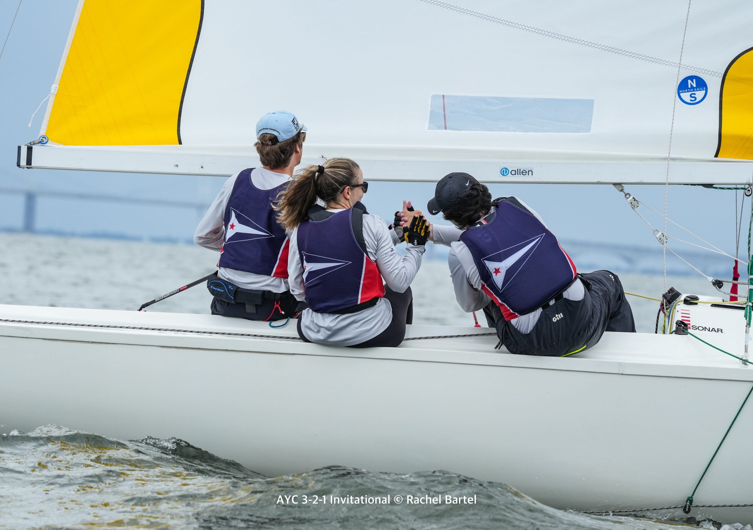 Three sailors wearing matching vests are sitting on the edge of a white sailboat, handling ropes and working together on the water during a sailing competition.