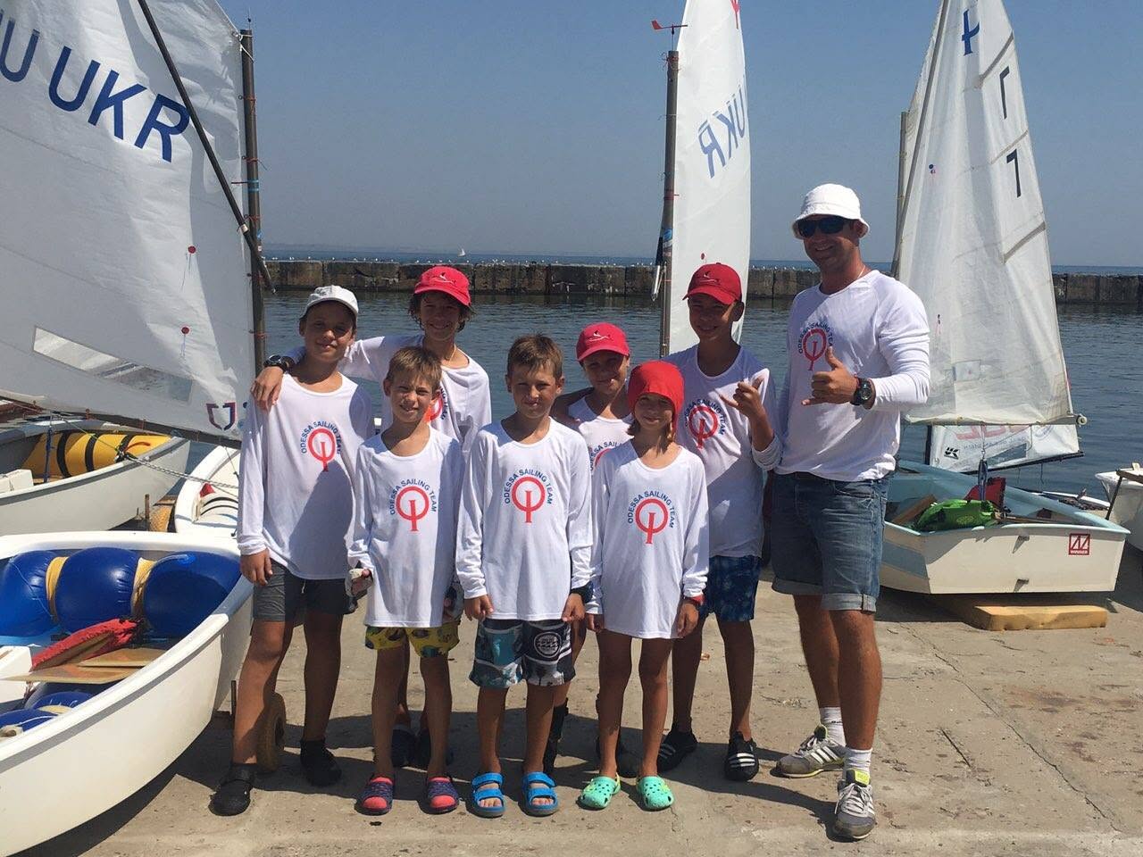 A group of children and an adult, all in matching white shirts and caps, stand together in front of small sailboats at a marina on a sunny day.