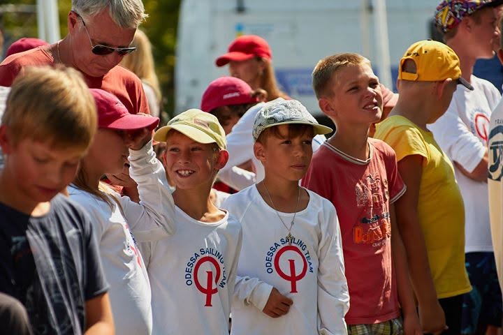 A group of children wearing “Odessa Sailing” shirts and hats stand outside together, some smiling, with adults and more kids in the background on a sunny day.