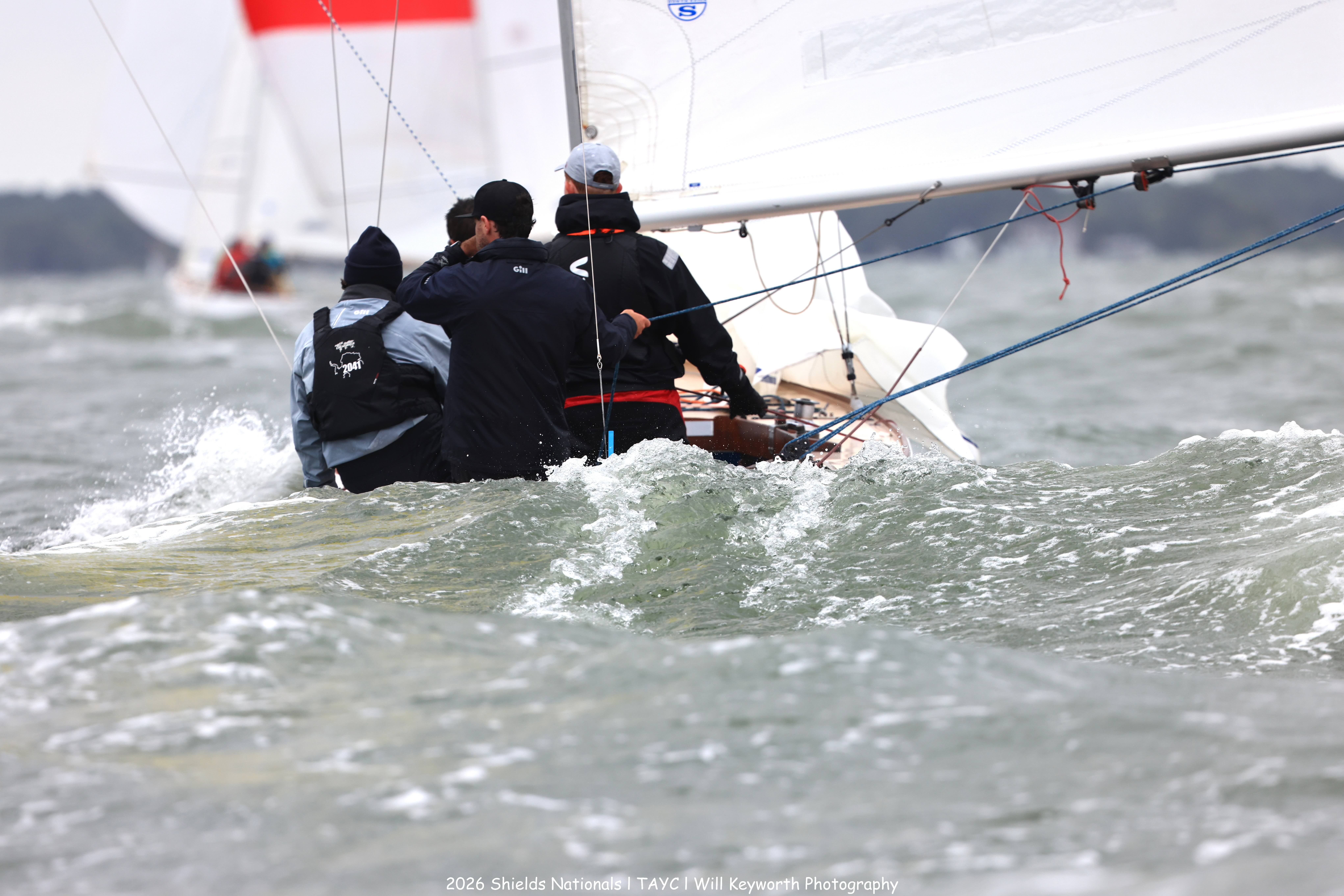 Three people sailing a small boat in choppy water, viewed from behind. Other sailboats are visible in the distance on a cloudy day.