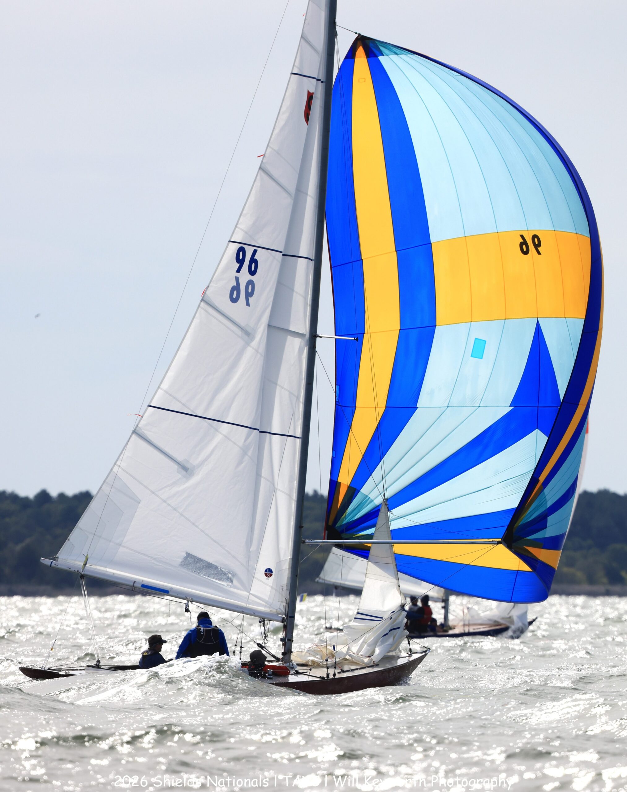 Two sailboats race on choppy water; one has a white sail marked “96” and the other displays a colorful blue and yellow spinnaker. The shore and trees are visible in the distance under a cloudy sky.
