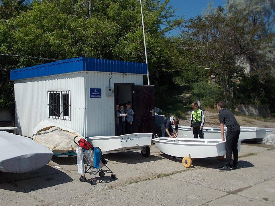 Children stand near small white boats outside a blue-and-white building, with trees in the background. Some kids are looking inside a boat while others watch from the doorway.