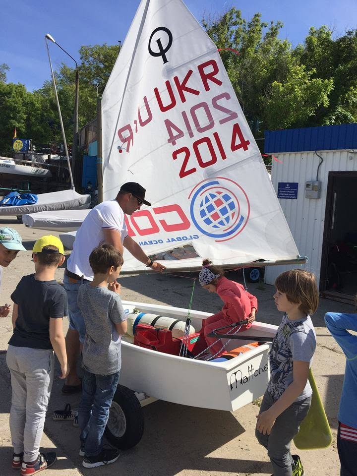 A group of children gathers around an adult who is showing them a small sailboat on a cart. The sail has “UKR 2014” written on it. Trees and buildings are visible in the background.