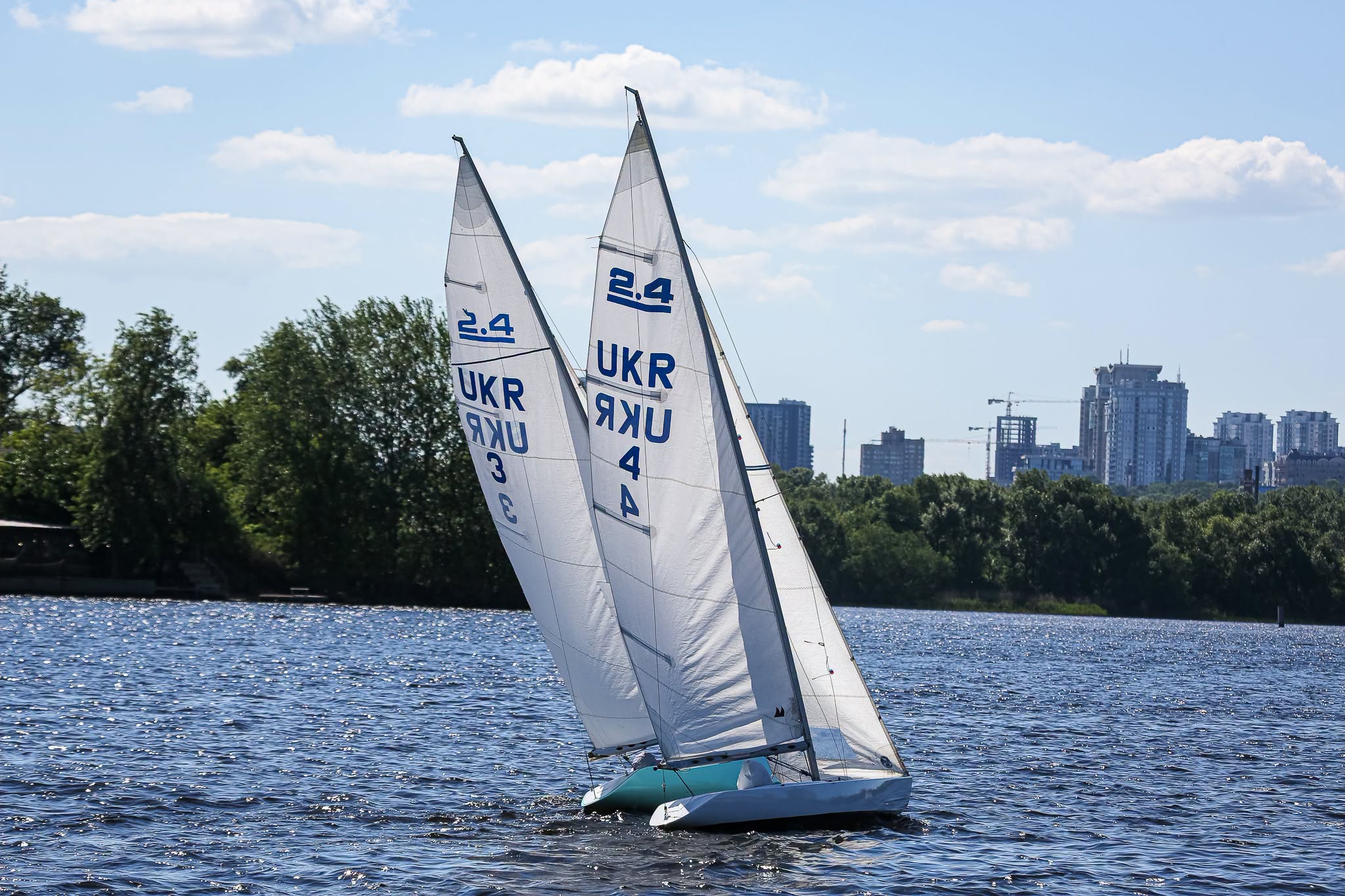Two small sailboats with white sails and blue “UKR” markings race on a river, with green trees and tall city buildings in the background under a partly cloudy sky.
