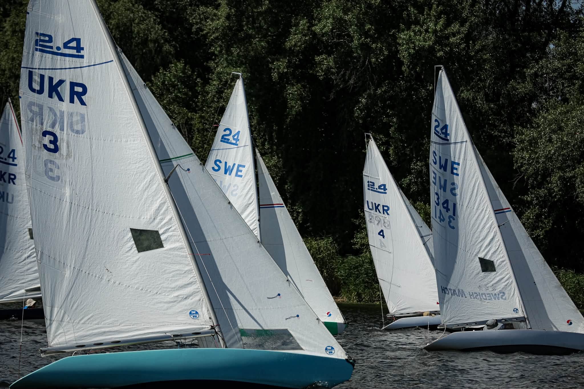 Several small sailboats with white sails, marked with country codes “UKR” and “SWE,” race on a body of water near a tree-lined shore.