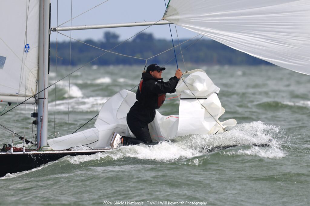 A person in black sailing gear and cap manages a large, white sail on a small sailboat in choppy waters. Waves splash against the boat, and a forested shoreline is visible in the background.