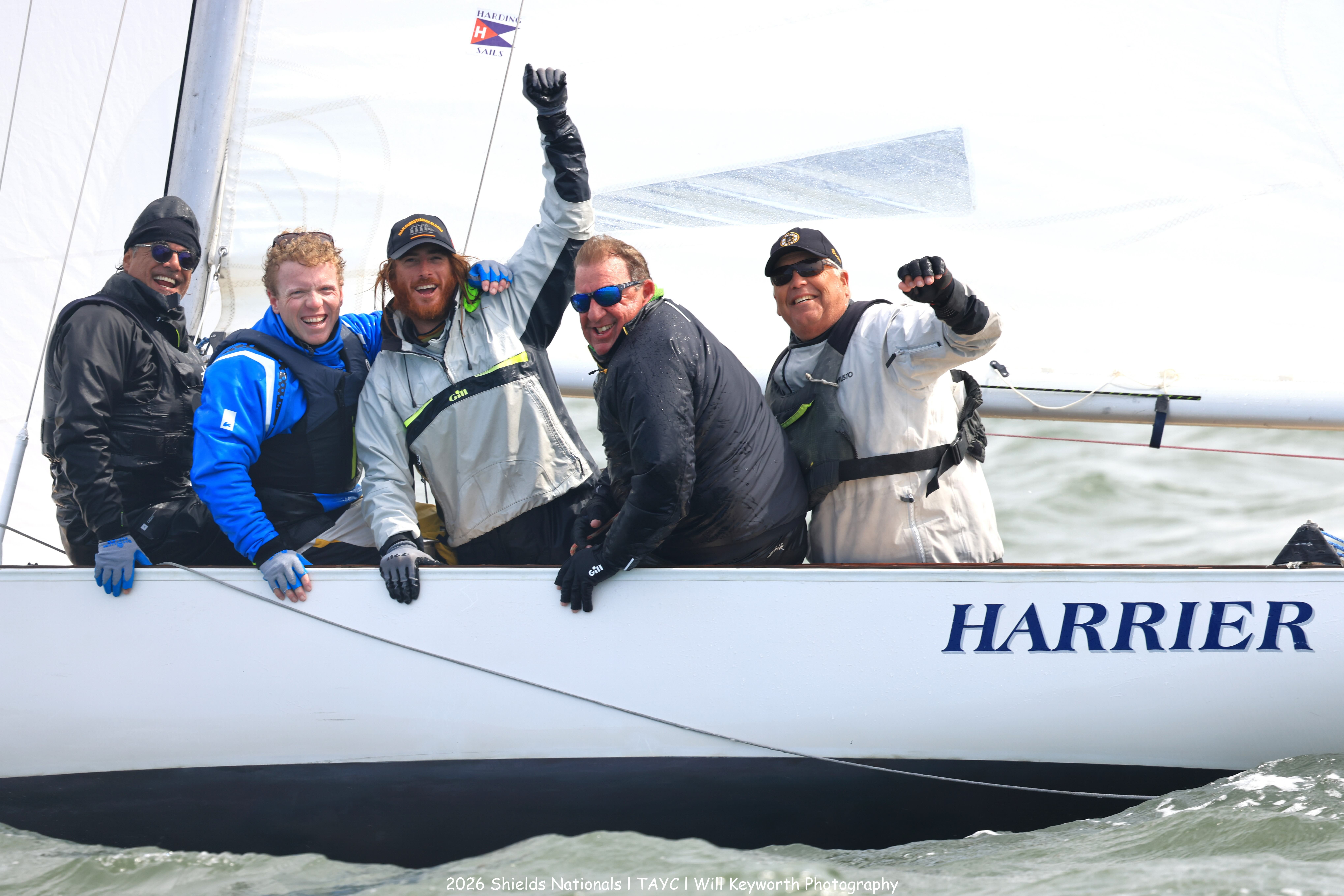 Five smiling people in sailing gear pose on a sailboat named “HARRIER” on the water, raising their hands in celebration. The weather appears windy and cool.