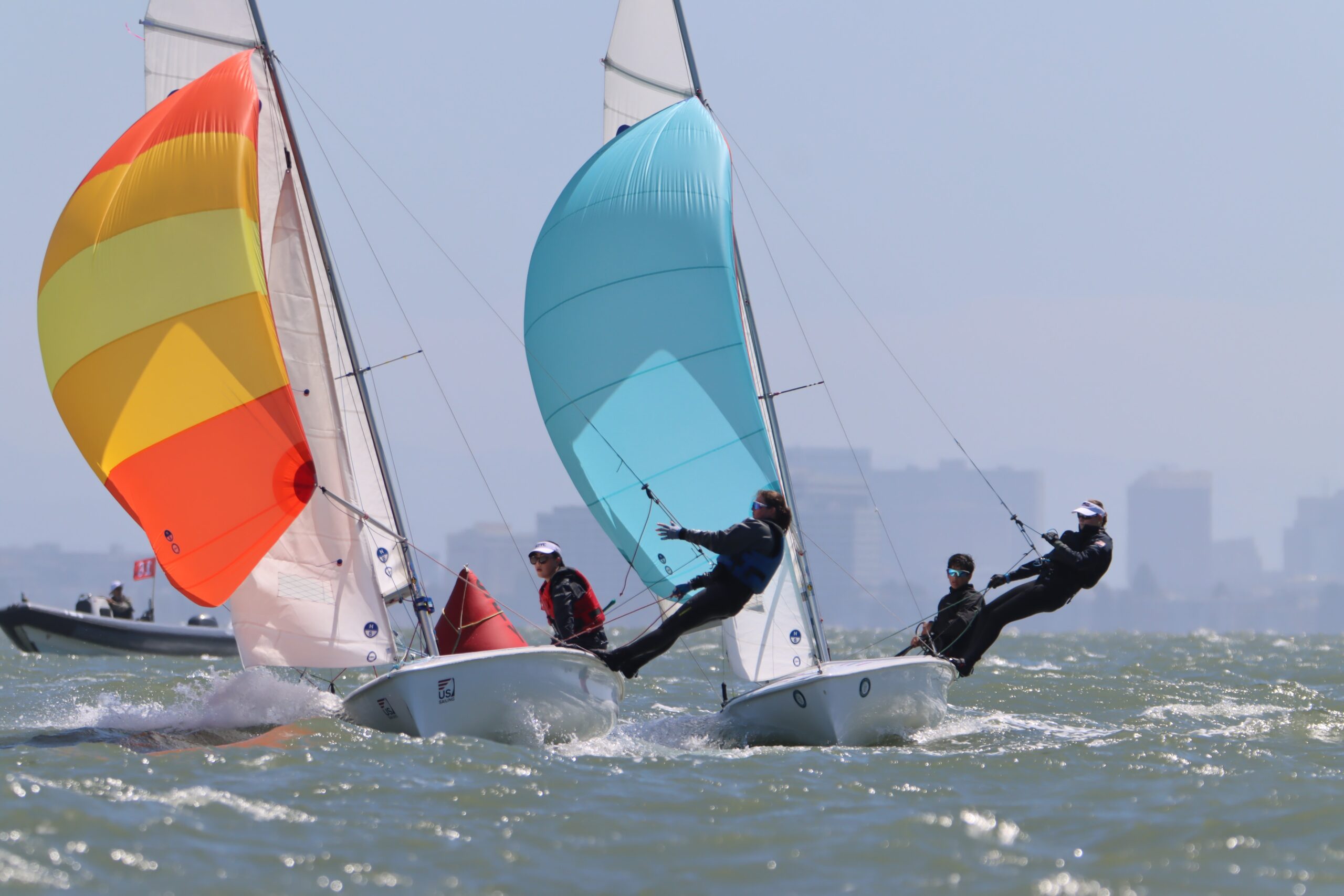 Three small sailboats race on choppy water, each with a colorful spinnaker sail. Sailors lean out over the edge for balance, and a city skyline is visible in the hazy background.