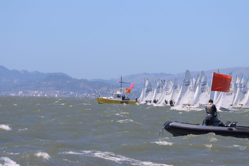 Sailboats line up for a race near a yellow official boat and a small boat with an orange flag on choppy water, with hills and a city visible in the distant background.