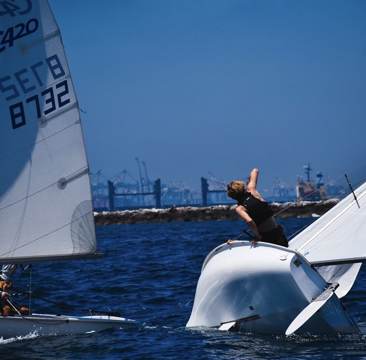 Two small sailboats on the water; one is capsizing as a person tries to balance on its side. The other boat sails nearby. Industrial cranes are visible in the background. The sky is clear and blue.