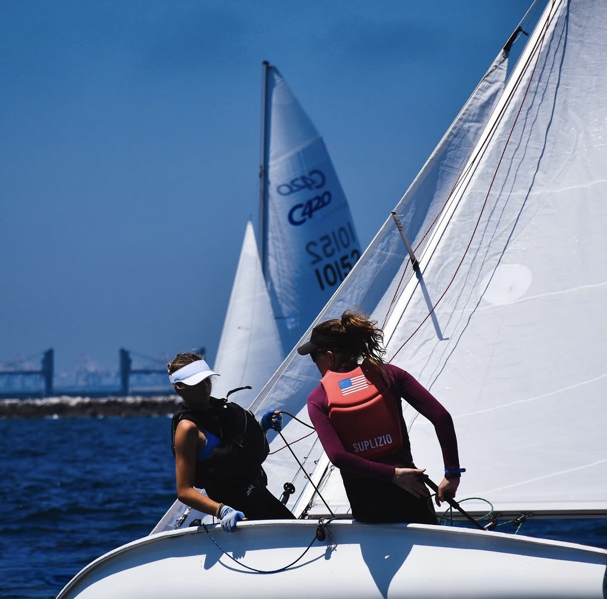 Two women sailing a boat with white sails on the ocean, one wearing a white visor and the other a red vest with a U.S. flag. A bridge and another sailboat are visible in the distance under a clear blue sky.