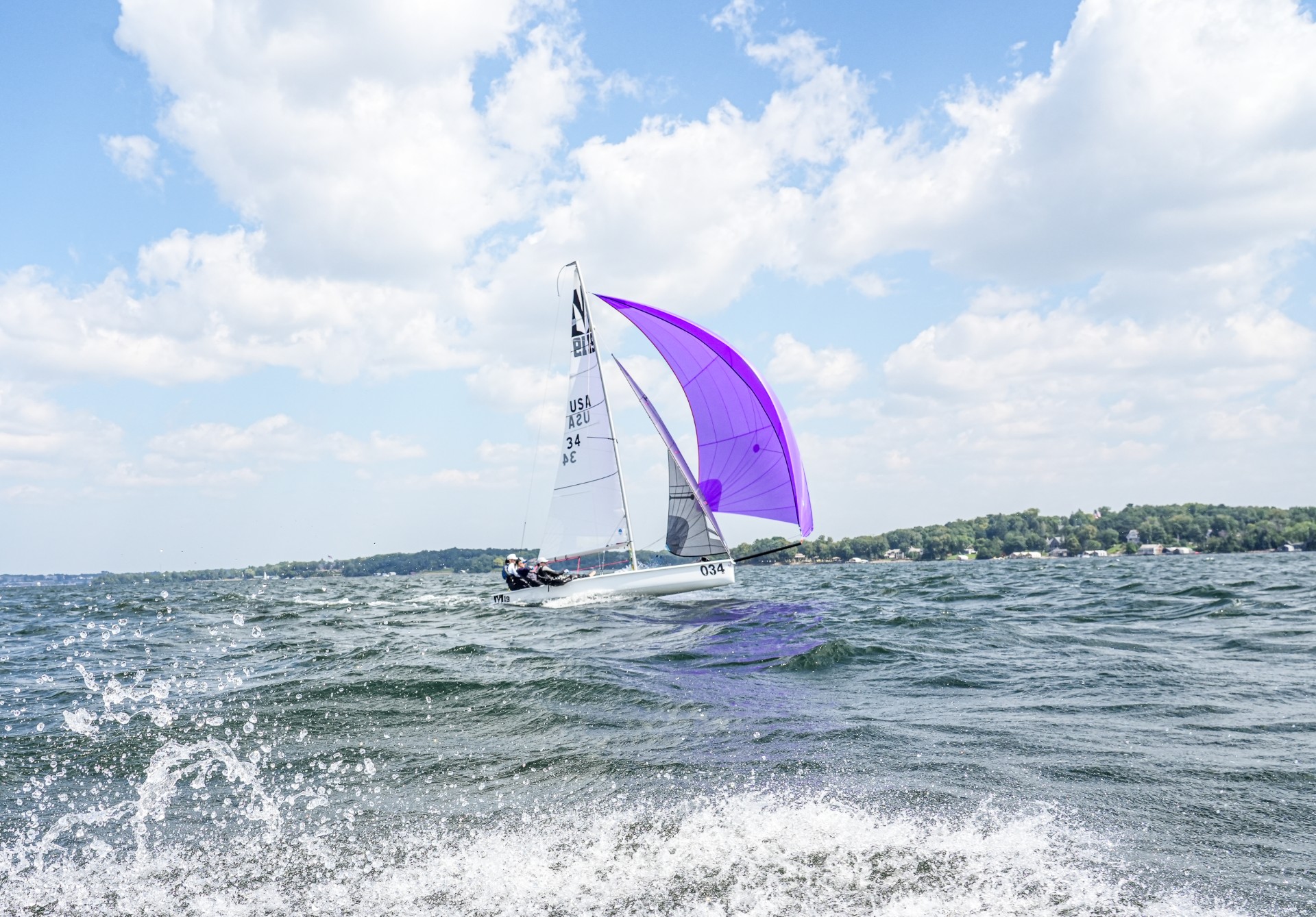 A sailboat with a bright purple sail glides on choppy water under a partly cloudy sky, with a distant treeline and shore visible in the background.