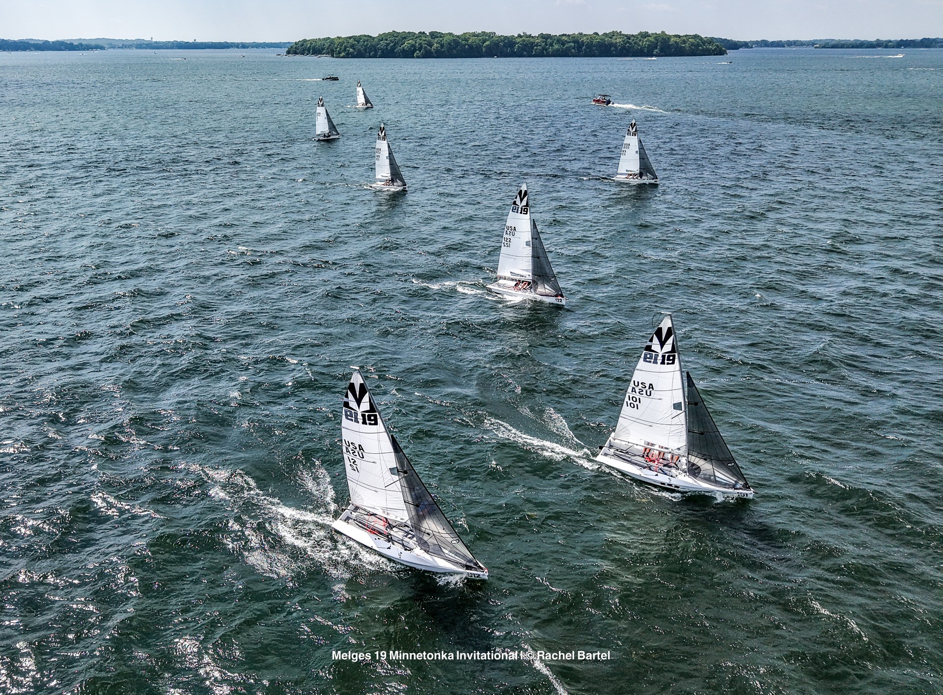 A group of sailboats race across a large, choppy lake under a clear sky, with a tree-covered island in the distance. The boats have white sails marked with “USA” and other numbers.