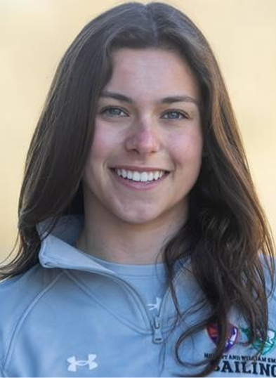 A young woman with long brown hair smiles at the camera, wearing a light gray zip-up jacket with a logo and the word “SAILING” partially visible. The background is blurred and neutral-colored.