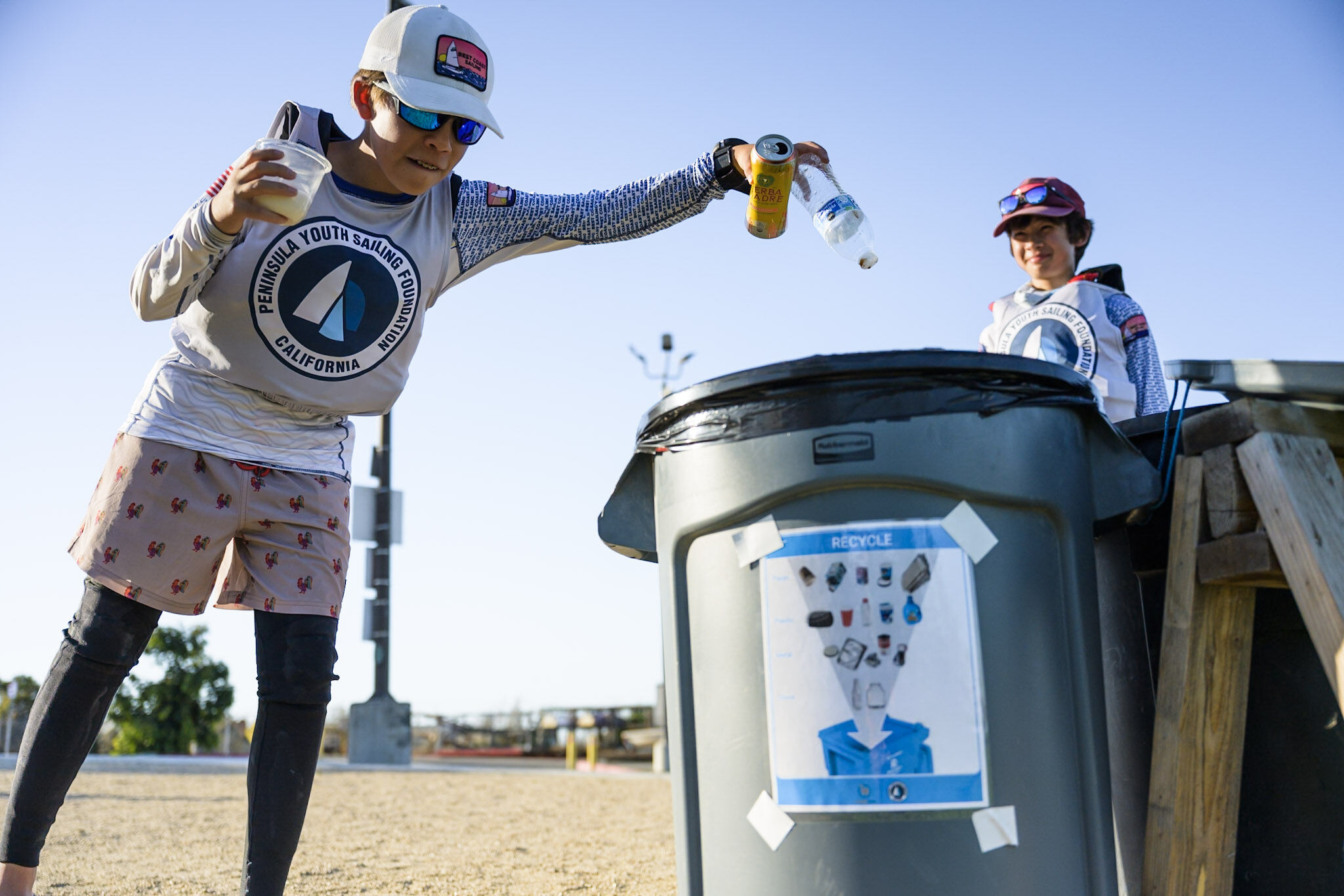 A young sailor wearing a branded performance shirt tosses a plastic bottle and a soda can into a recycling bin.