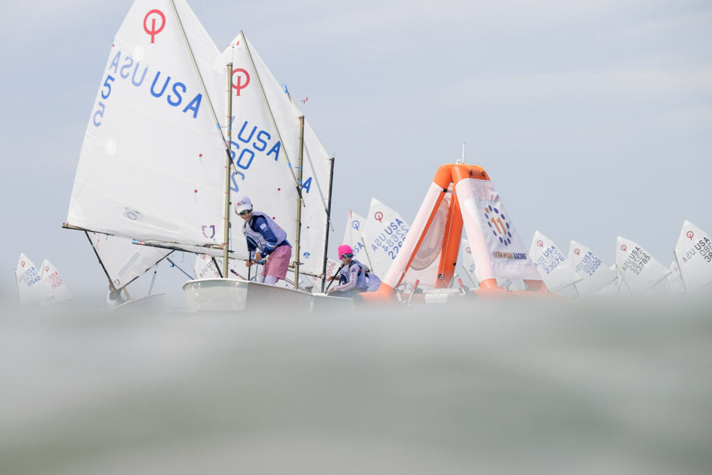 Two young sailors in small sailboats navigate past an orange inflatable buoy during a race.