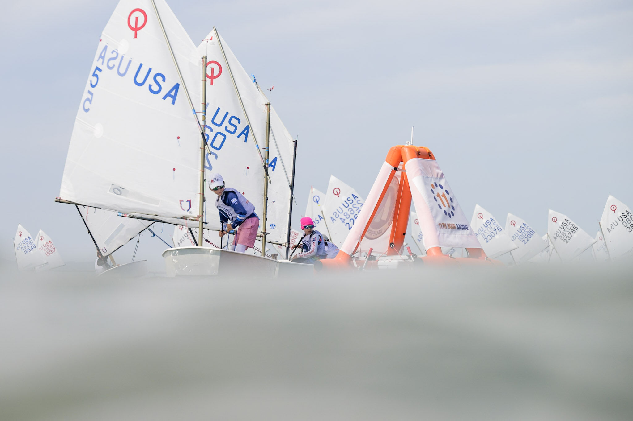 Two young sailors in small sailboats navigate past an orange inflatable buoy during a race.