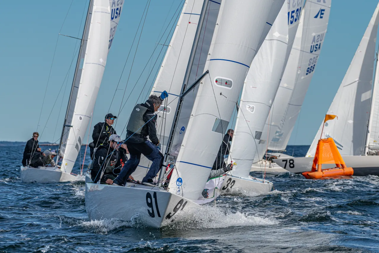 Several sailboats racing on the water, with crew members actively adjusting sails and lines on the deck of the lead boat numbered 91.
