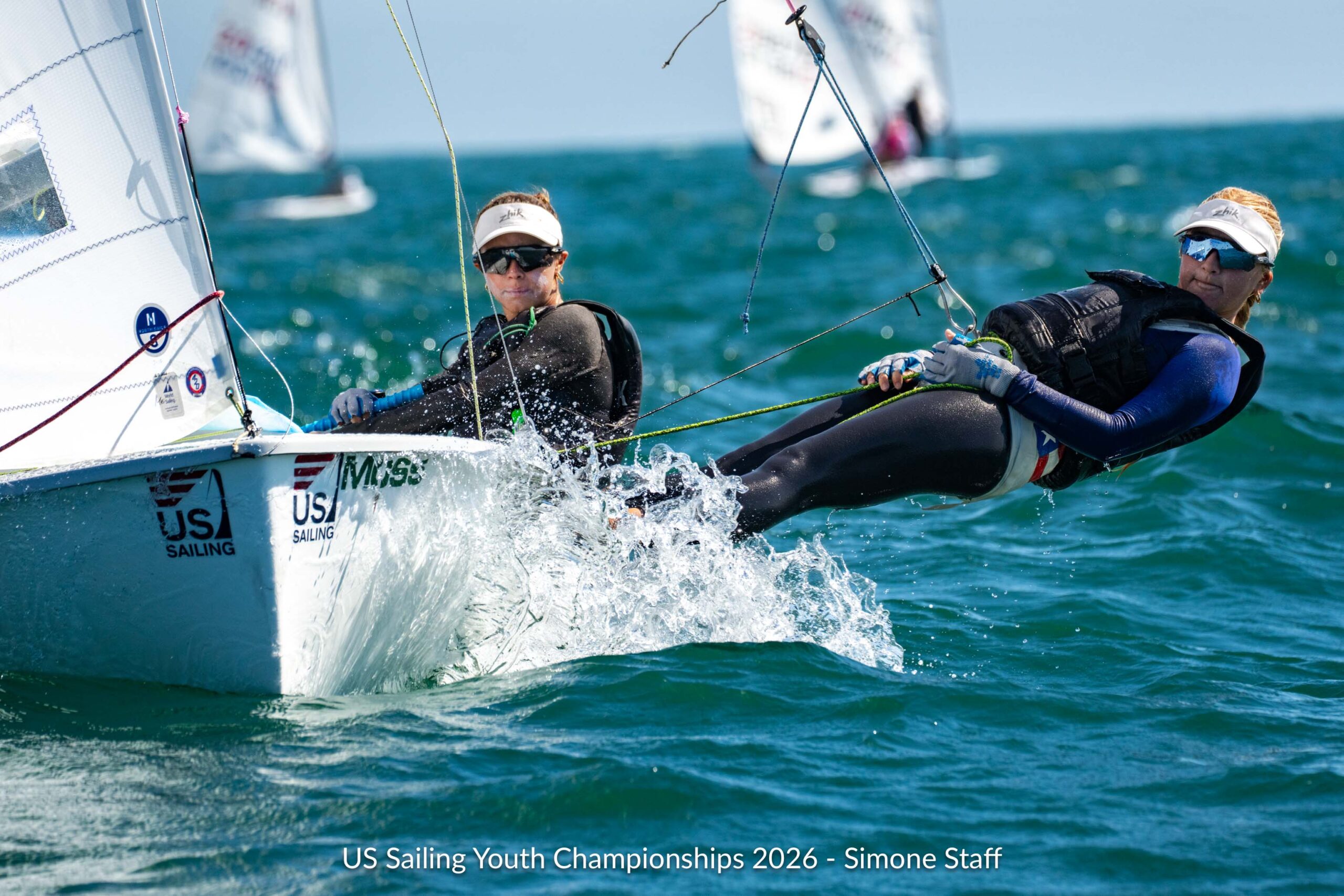 Two sailors in wetsuits and visors navigate a small sailboat through choppy water during the US Sailing Youth Championships.