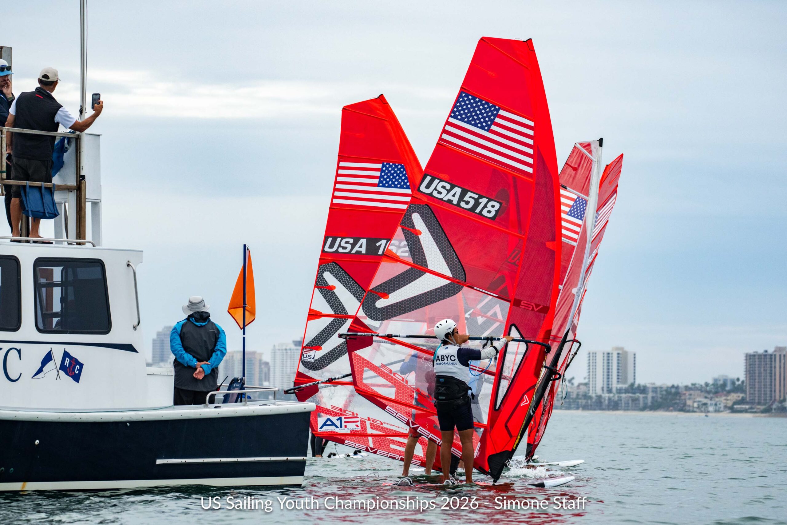 Windsurfers with red sails featuring the US flag and boat numbers gather near a support boat at the US Sailing Youth Championships.