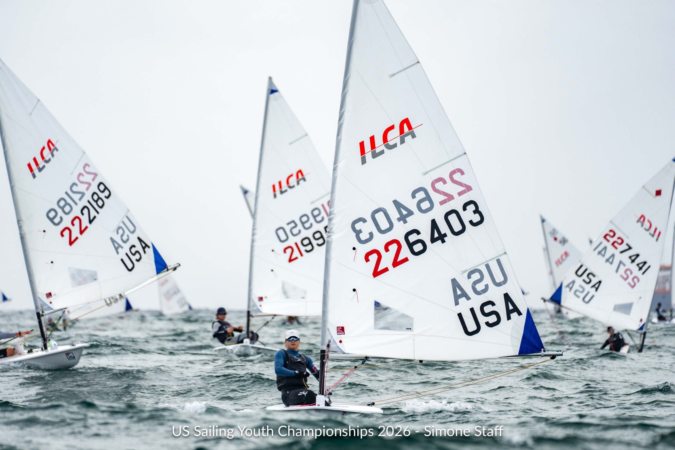 Several ILCA sailboats racing on the water, including a foreground sailor wearing a blue long-sleeve shirt and cap.